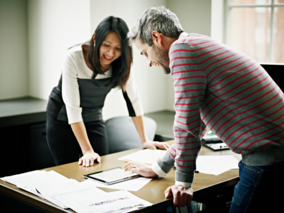 A woman and man looking at a tablet amidst paperwork on a desk together.