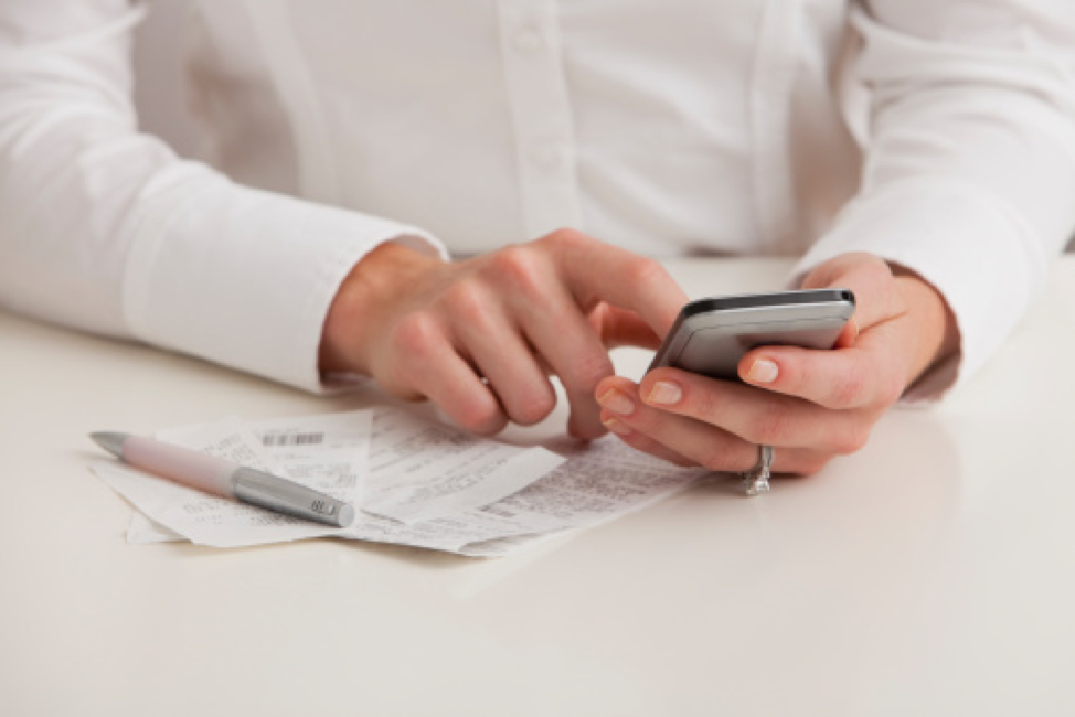 Hands typing on a cell phone above a pile of receipts on a desk.