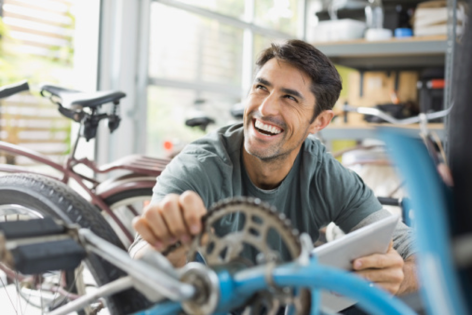 Man joyously smiling upward while sitting among multiple bikes. He's holding a tablet and bike part.