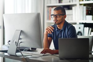 Man holding pen to chin while looking at computer monitor