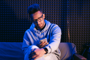 Curly man wearing glasses hold smartphone while sitting on couch
