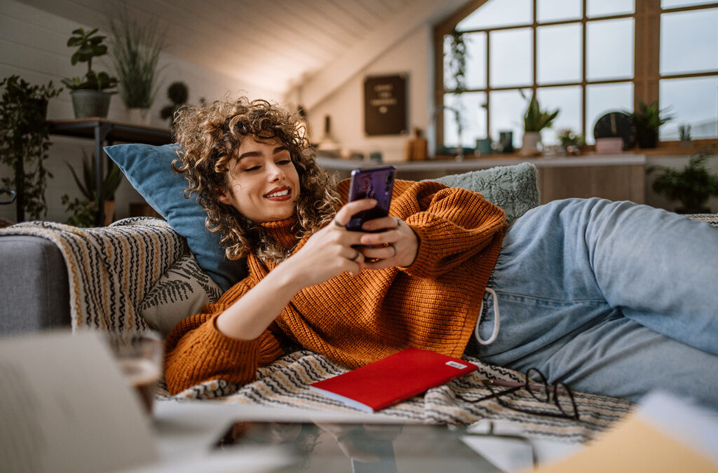 A smiling woman is lying on her couch looking at her phone.
